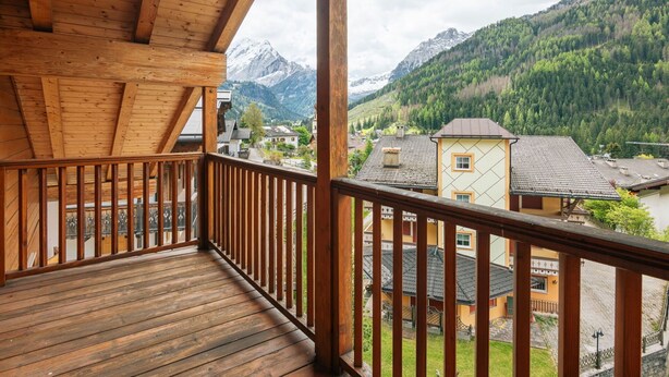 A wooden balcony with a view of a mountain village and snow-capped peaks.