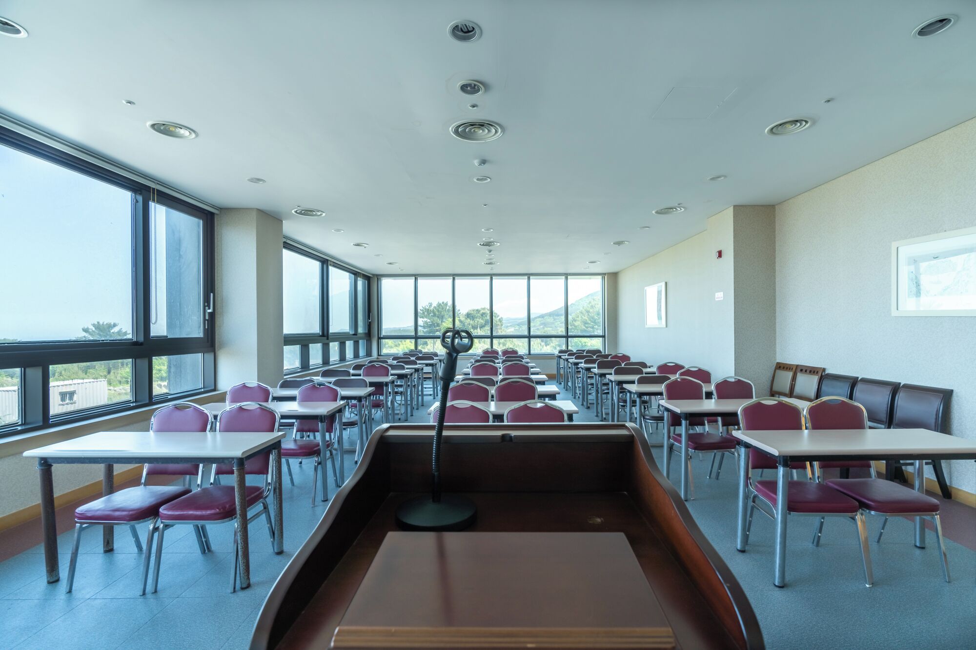 Une salle de classe avec des rangées de bureaux et de chaises, de grandes fenêtres et une allée centrale.