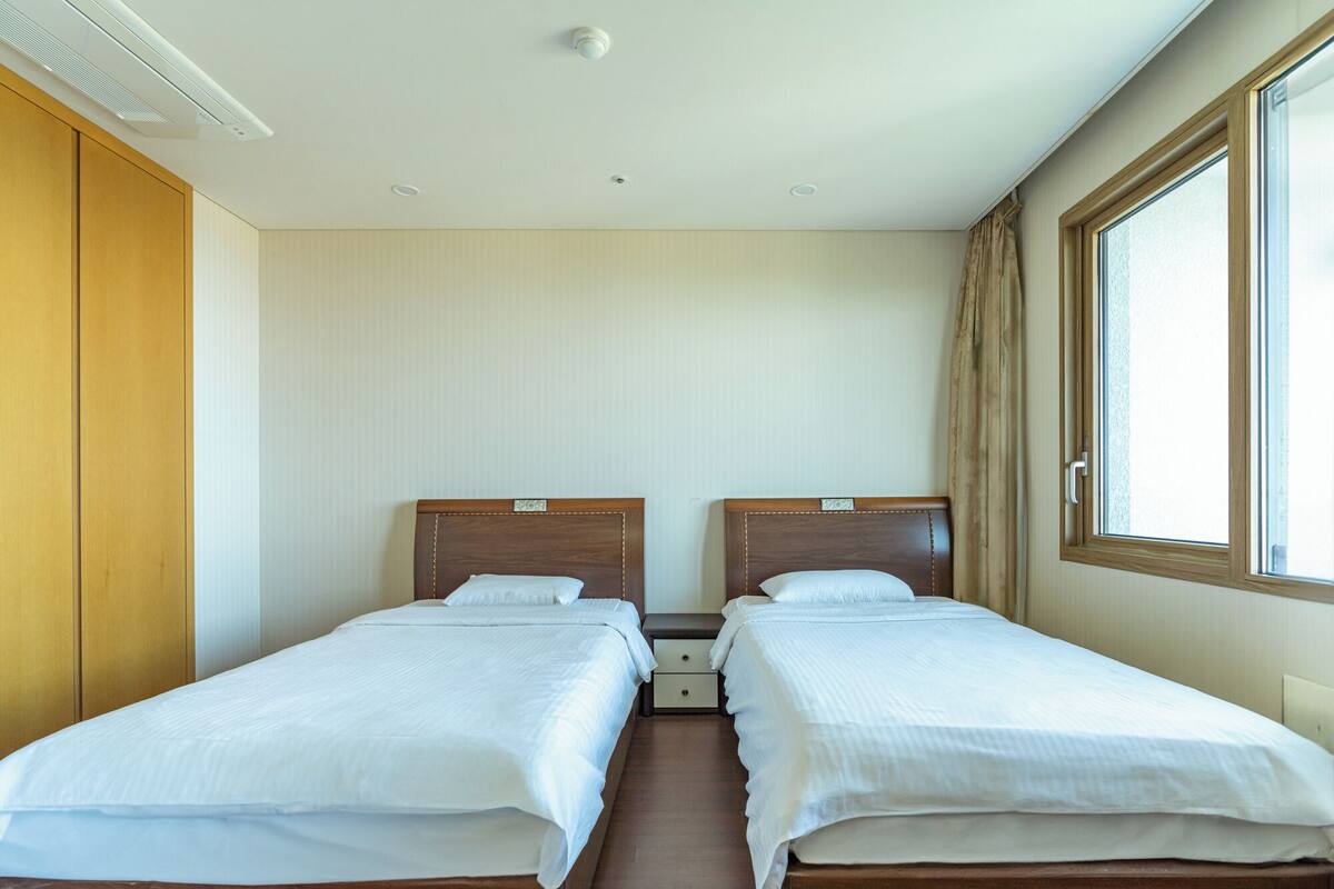Una habitación de hotel con dos camas individuales, cabeceros de madera, una ventana con cortinas e iluminación empotrada en el techo.