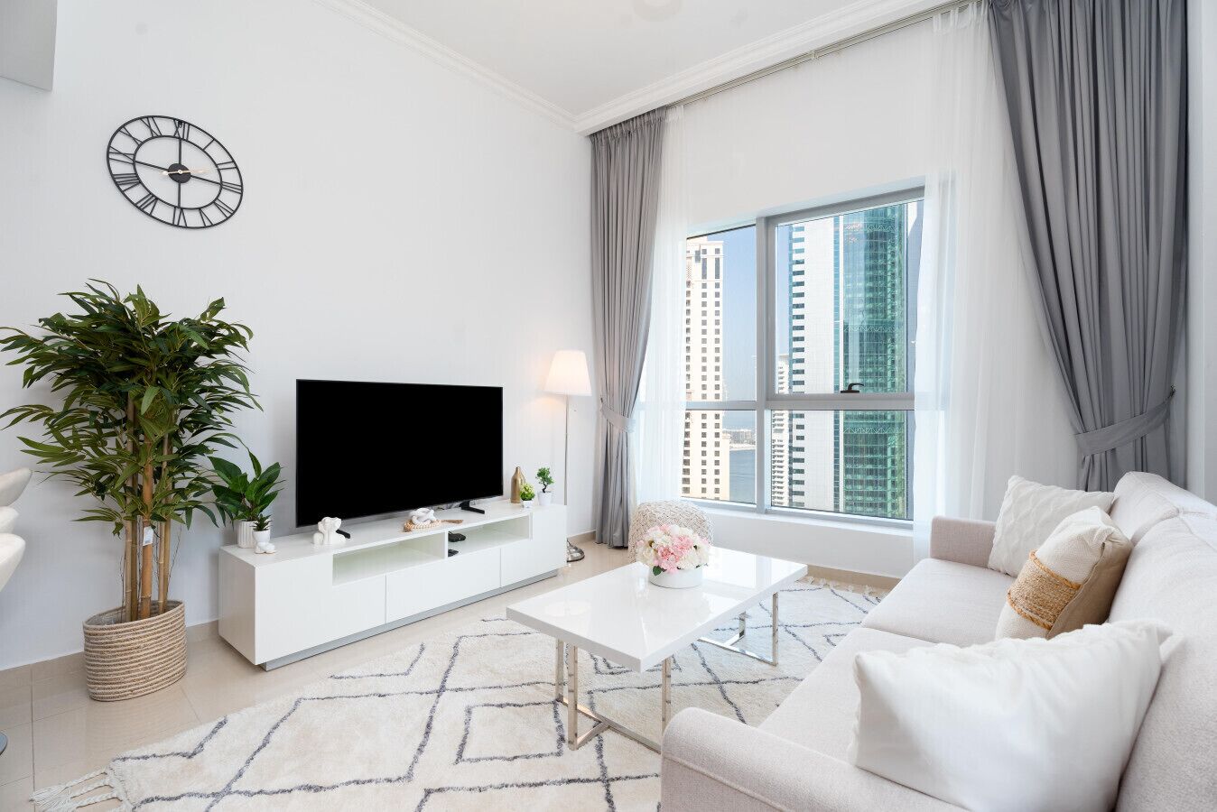 Una sala de estar moderna con un televisor de pantalla plana, una planta grande en maceta, un sofá blanco y vistas de edificios altos a través de un ventanal.