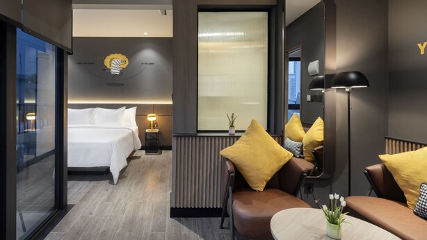 A modern hotel room with a bed, a seating area with yellow cushions, a mirror, and a small table with a plant.