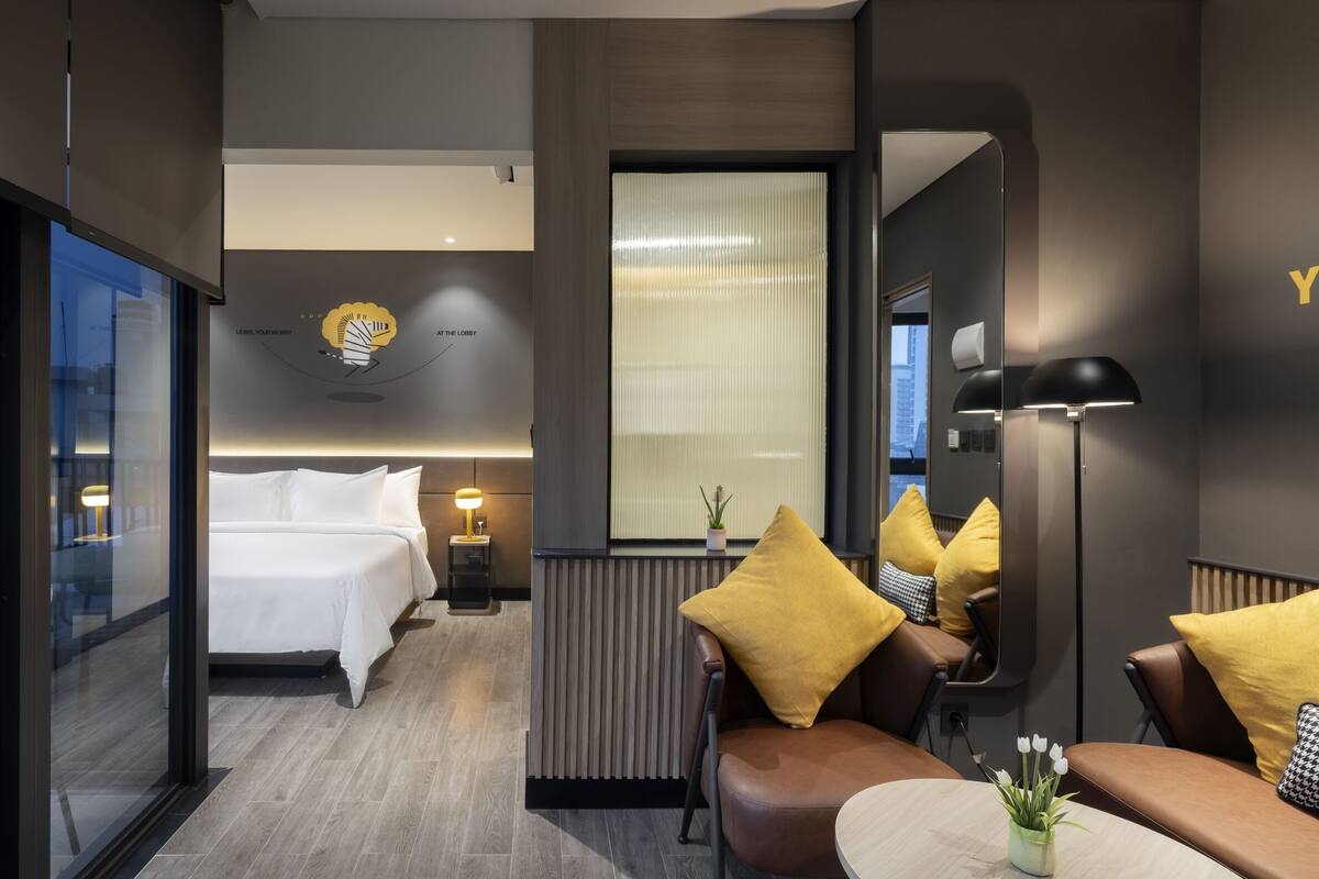 Una habitación de hotel moderna con una cama, un rincón de estar con cojines amarillos, un espejo y una mesita con una planta.