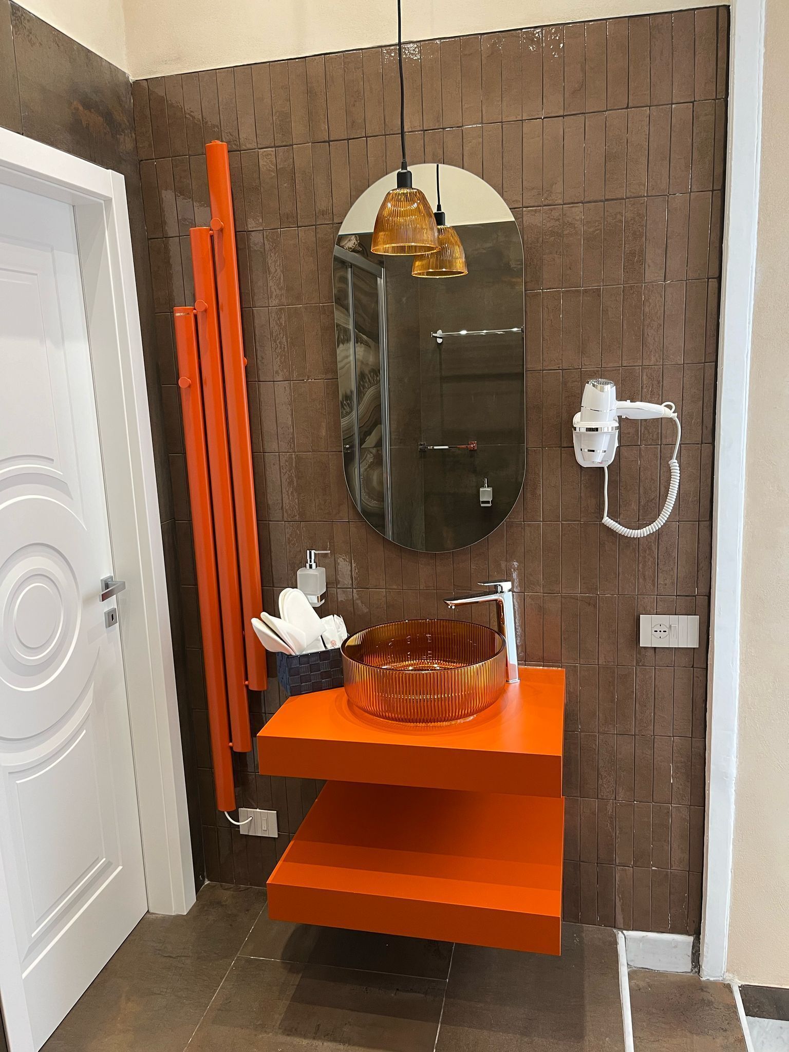 A bathroom with a brown tiled wall, a white door, a round mirror, a white hairdryer, an orange sink, and a towel rack.