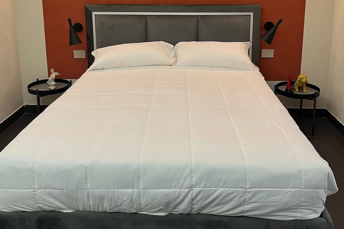 A neatly made bed with a grey headboard and white bedding, flanked by two nightstands with lamps and a small table with a figurine.