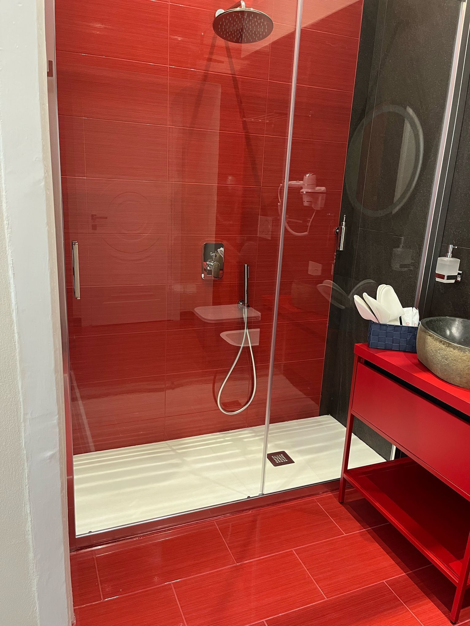 A modern bathroom with a red tiled shower enclosure, a glass door, a white sink, and a red vanity with a bowl.