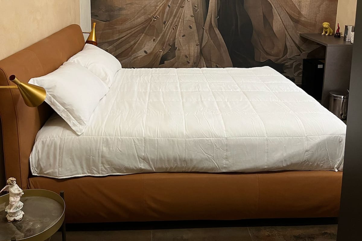 A bedroom with a bed, a brown headboard, a white pillow, a small table with a statue, and a mural on the wall.