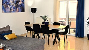 A modern living room with a sofa, a dining table with chairs, a painting on the wall, and a view through a window. - BH007 - A - Alerine Home (Heraklion)