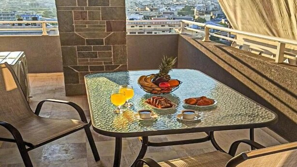 A glass-top table with breakfast items, a fruit bowl, and a drink on a balcony overlooking a cityscape. - BH007 - A - Alerine Home (Heraklion)