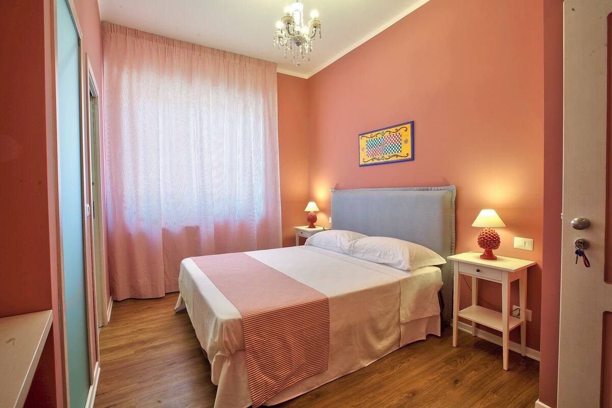 A bedroom with a bed, two bedside tables with lamps, a chandelier, a door with keys, and a colorful artwork on the wall.