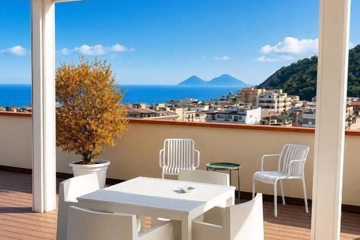 A covered outdoor seating area with a table and chairs, a potted plant, and a view of the sea and buildings.
