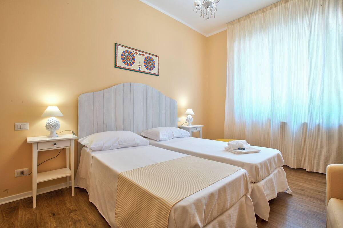 A hotel room with two beds, a chandelier, a painting on the wall, and a window with curtains.