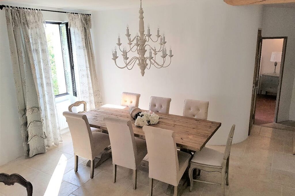 Une salle à manger avec une table en bois et six chaises, un lustre et une fenêtre avec des rideaux.