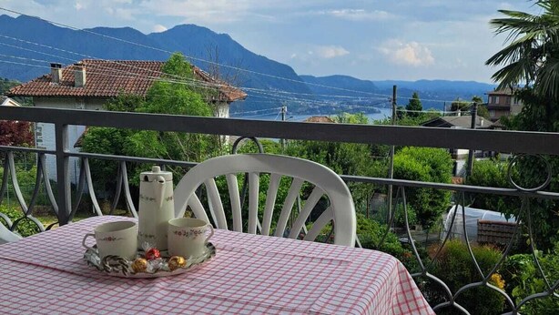 A table set with a checkered tablecloth, white chairs, and a tray with pastries and cups, overlooking a mountainous landscape. - RED FOUNTAIN HOUSE 2 (arizzano)