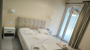 A neatly made bed with pillows and towels, a bedside table with a lamp, and a window with curtains. - Aggelika Apartments (Zakynthos)