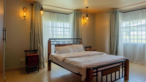 A bedroom with a wooden bed, two nightstands, a wall-mounted light fixture, and a window with curtains. - Gatundu Peaceful Tea Farm Stay w/activities (Gatundu)
