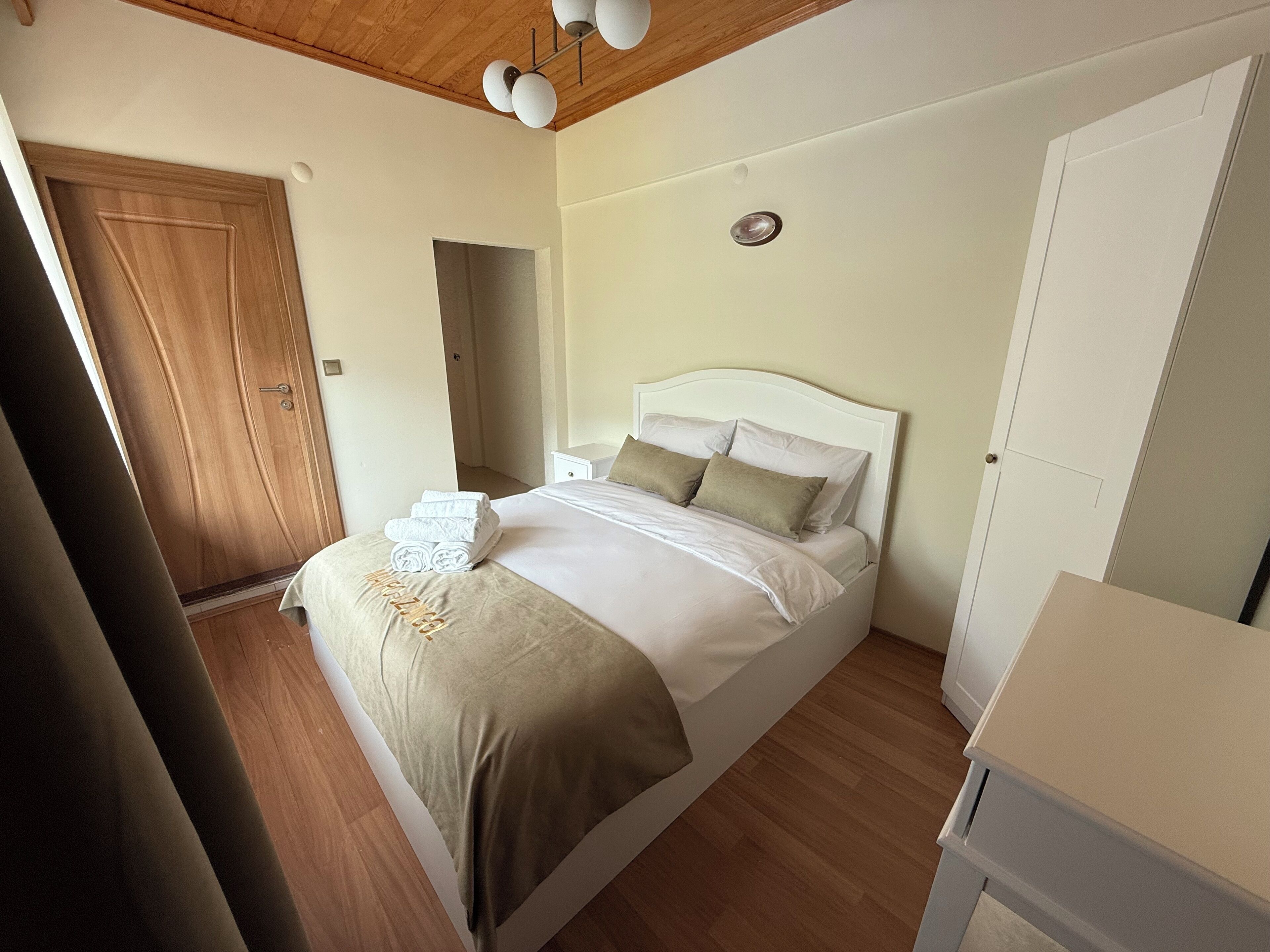 A bedroom with a bed, a wooden door, a white nightstand, and a ceiling light fixture.