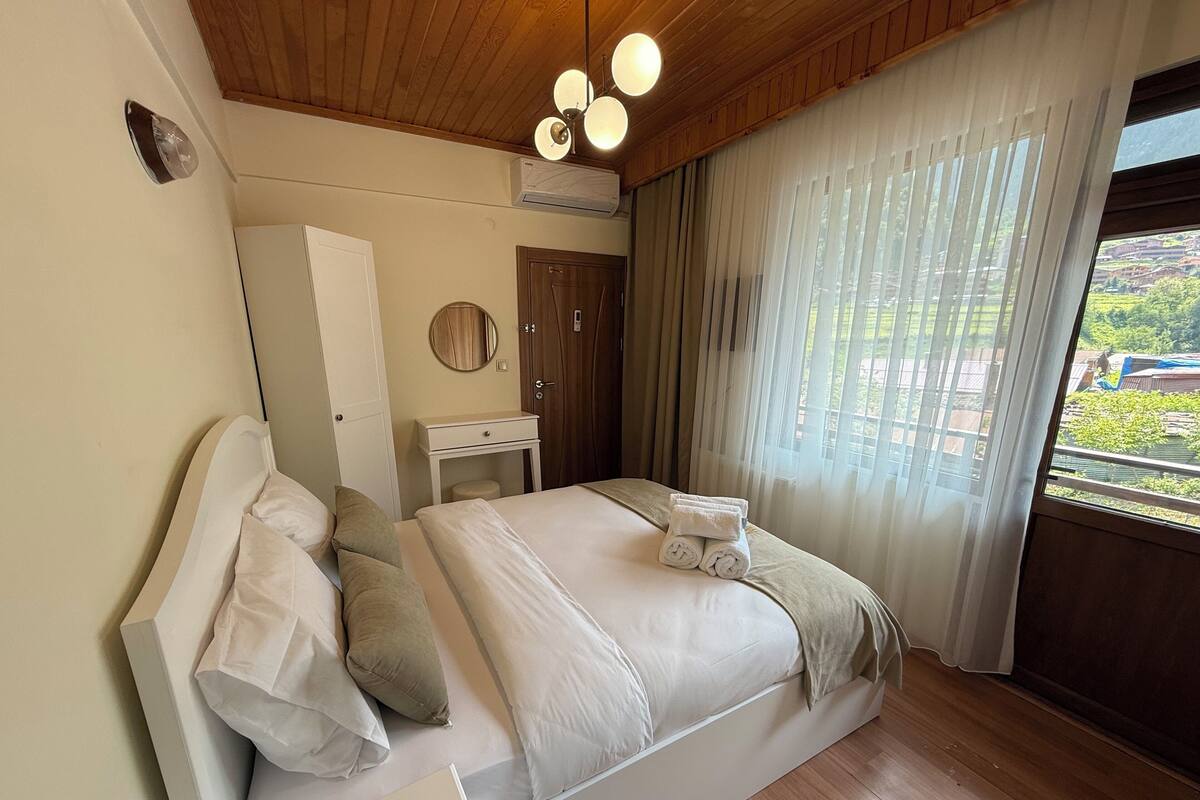 A bedroom with a wooden ceiling, a bed with white linens, a bedside table, a mirror, and a window with curtains.