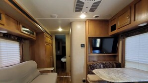 A compact RV interior with a sofa, a small table, a TV mounted on a cabinet, and a kitchenette area. - Serene Pinehurst Camper - Sleeps 2 - Pet Friendly (Magnolia)