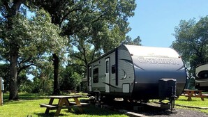 A stationary RV with a visible logo and text, parked on a grassy area with picnic tables nearby. - Serene Pinehurst Camper - Sleeps 2 - Pet Friendly (Magnolia)