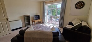 A living room with a sofa, a television on a wooden table, a dartboard on the wall, and a view of the outside through a large window. - Tidy, Spacious & Quiet 2BD on Edge of Cork City (Cork)