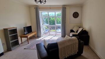 A living room with a sofa, a TV, a dartboard, and a window with curtains.