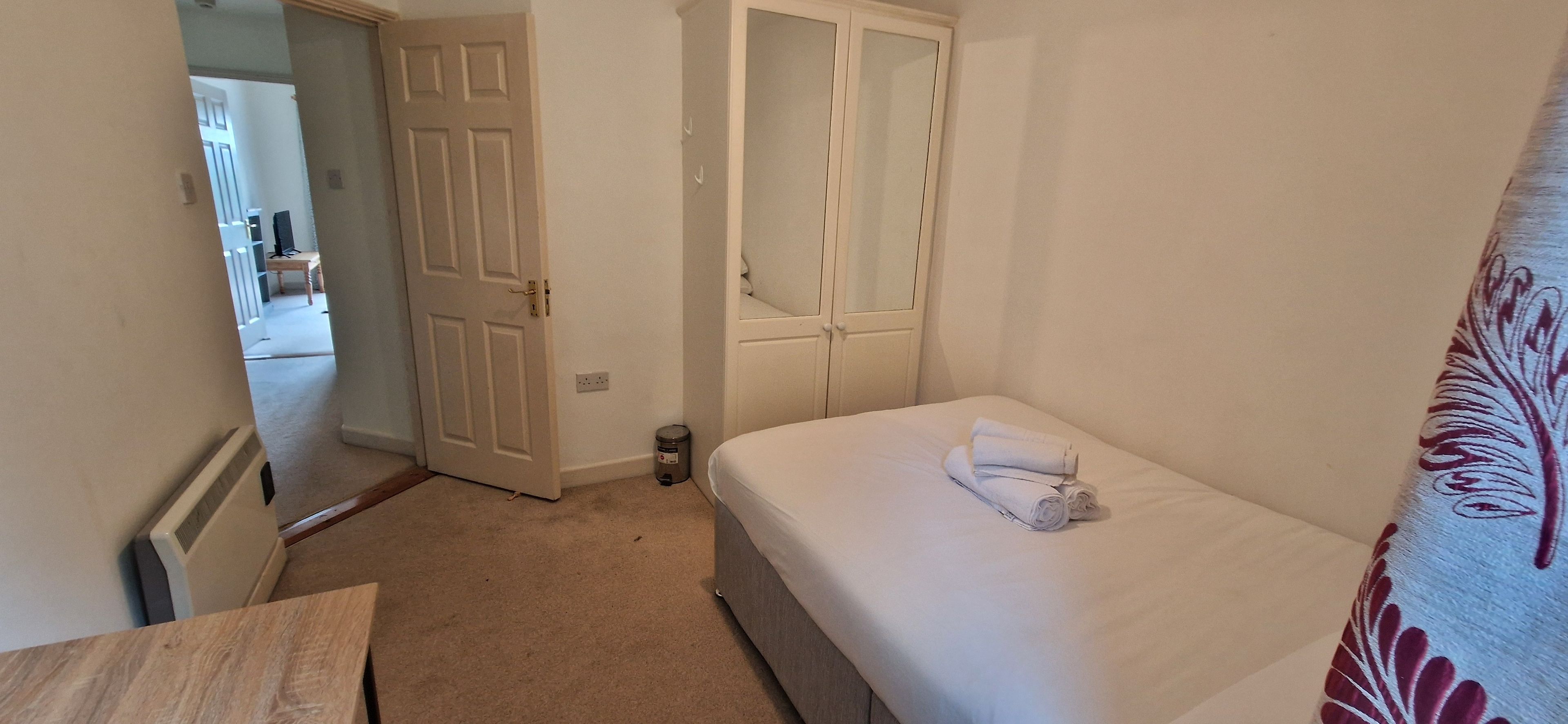 A bedroom with a bed, a bedside table, a door, and a small trash bin.