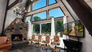 Une pièce spacieuse avec une cheminée en pierre, de grandes fenêtres et un coin salon confortable, comprenant des fauteuils et un canapé.