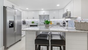 A modern kitchen with white cabinets, stainless steel appliances, a granite countertop, and a central island with bar stools. - Nautilus 2511 Gulf View 2 BR/2 BA 5th Floor 2026 REMODEL! (FORT WALTON BEACH)