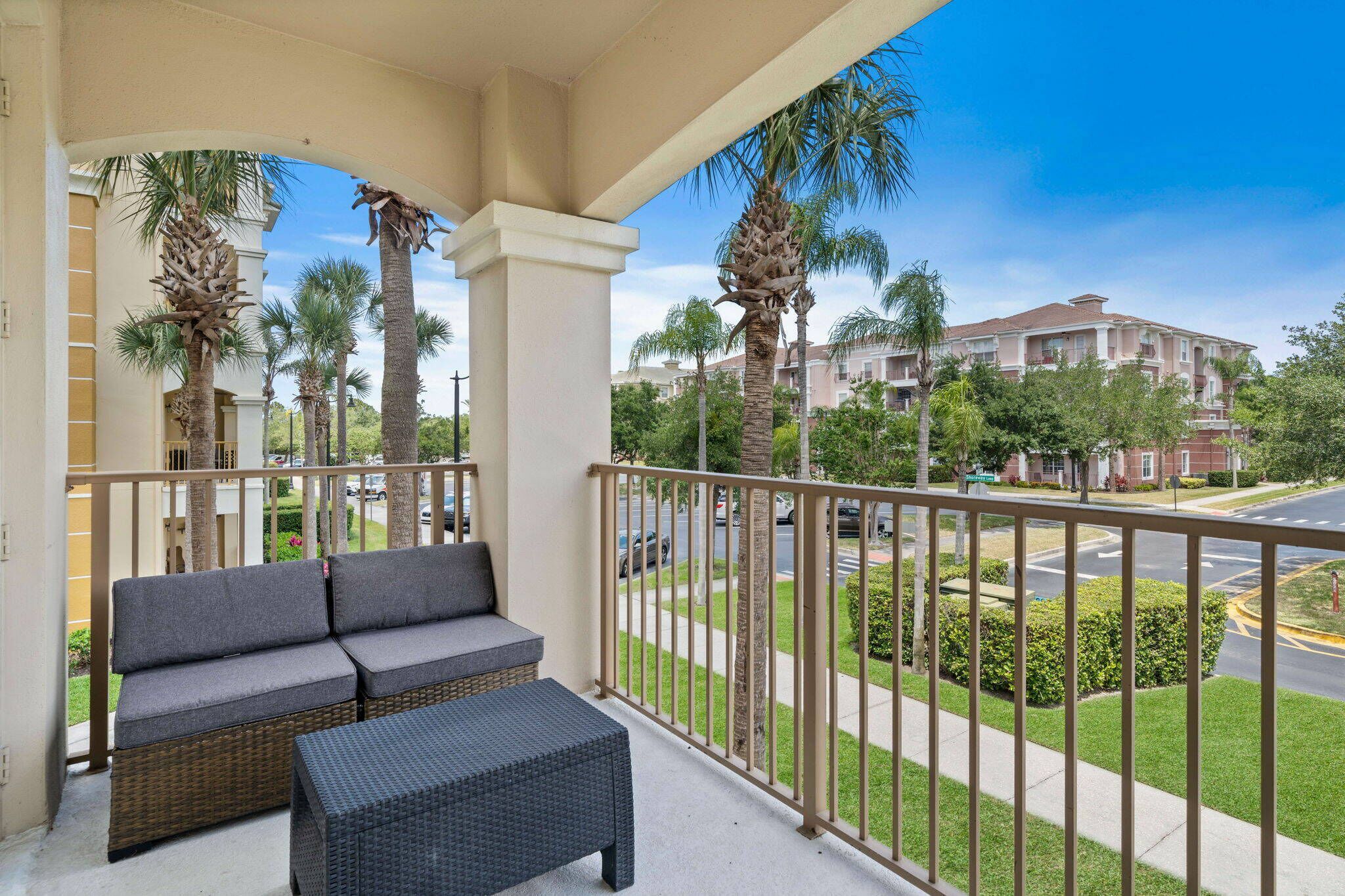 A balcony with a sofa and a small table, overlooking a street with palm trees and a building in the background.