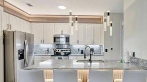 Una cocina moderna con armarios blancos, una isla central con asientos tipo barra, electrodomésticos de acero inoxidable e iluminación colgante.