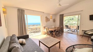 Ein geräumiges Wohnzimmer mit einer Couch, einem Couchtisch und einem Esstisch für vier Personen. Ein Balkon bietet Blick auf das Meer.