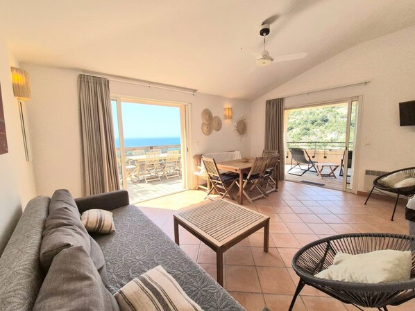 A spacious living area with a sofa, a coffee table, and a dining table set for four. There is a balcony with a view of the sea.