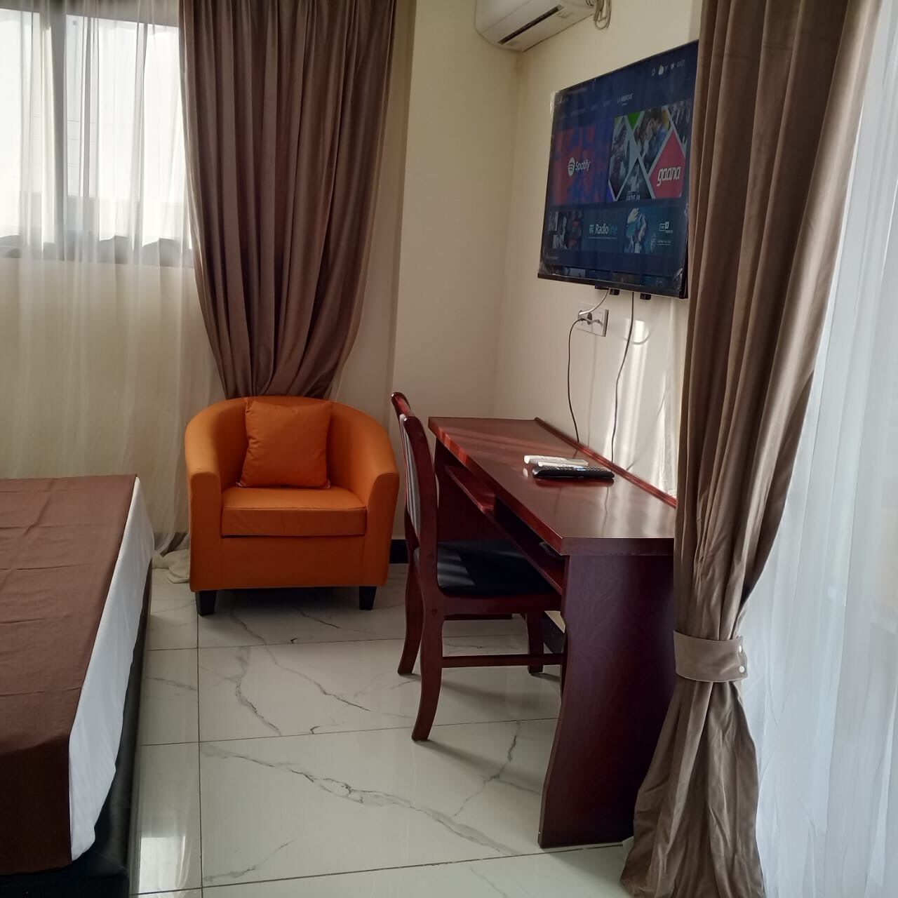 Ein Hotelzimmer mit einem Bett, einem orangefarbenen Sessel, einem Holztisch und einem an der Wand montierten Fernseher.