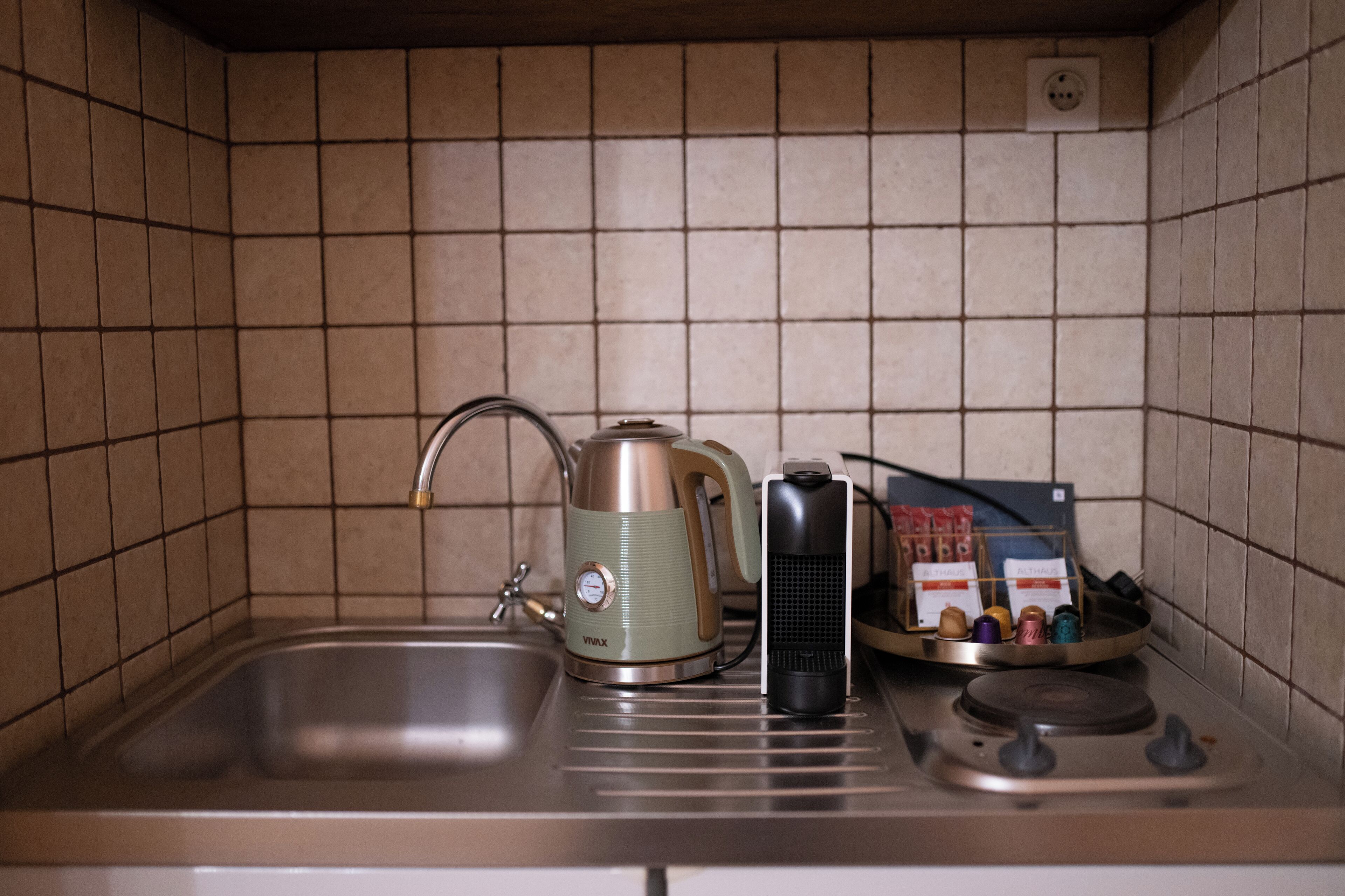 Una cocina con un lavamanos, una tetera, una cafetera y una bandeja con cápsulas de café.