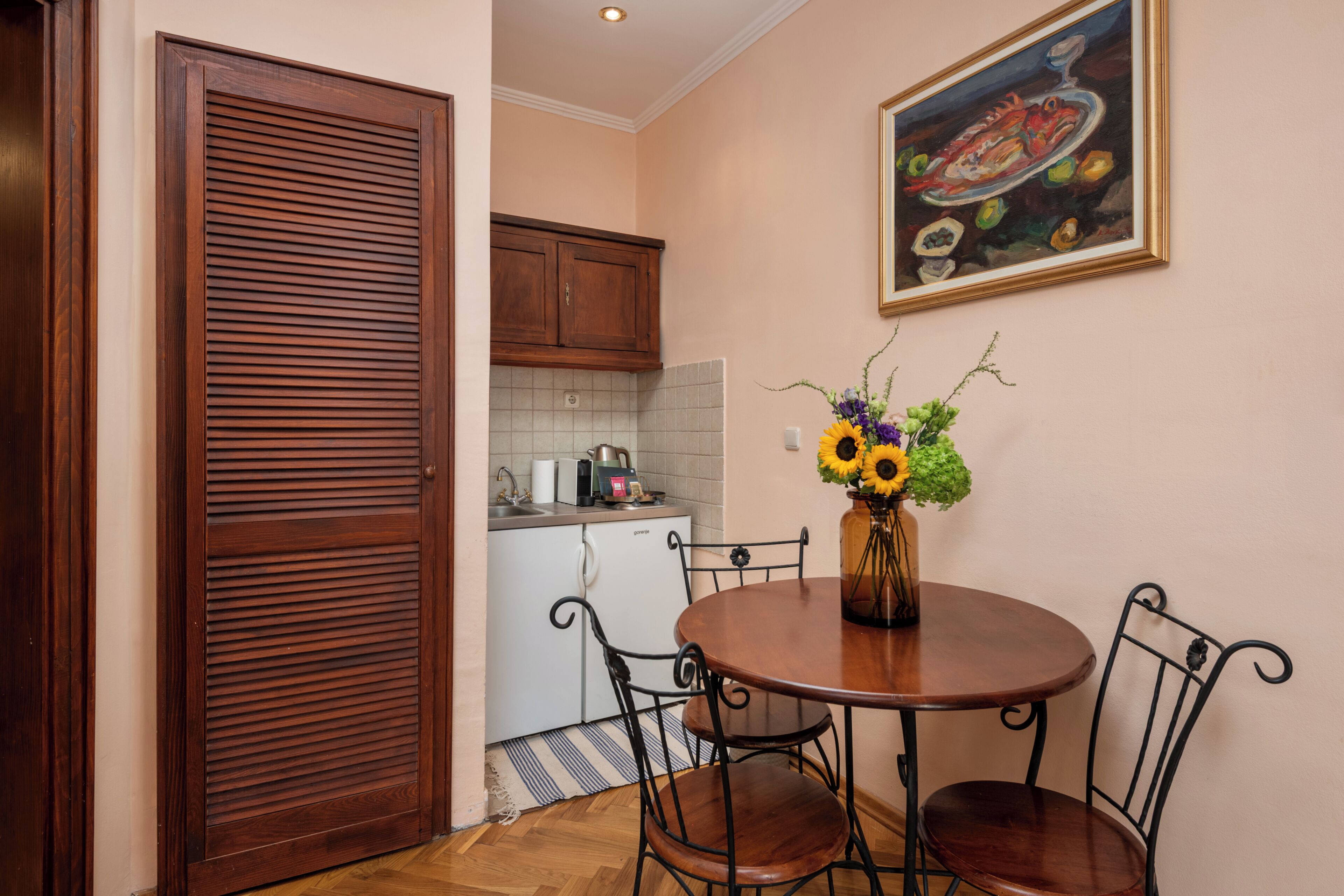 A dining area with a wooden table, four chairs, a vase with flowers, a painting on the wall, and a kitchen in the background.