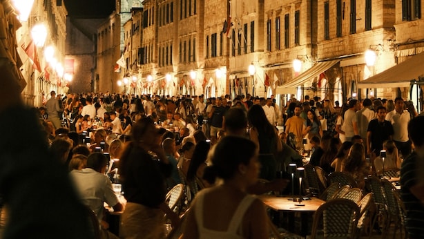 A crowded street scene at night with historic buildings and a tower in the background. - Antaeus Old Town Residence (Dubrovnik)