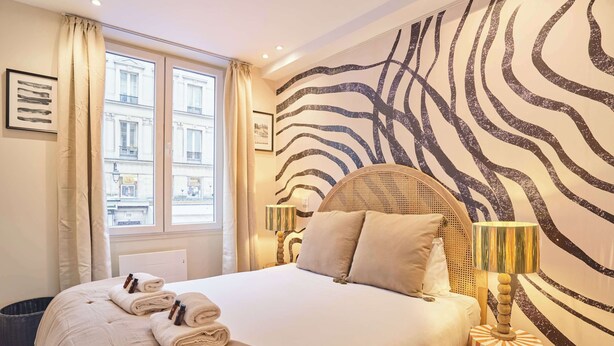 Una habitación de hotel con una cama grande, un ventanal con cortinas, una pared con estampado de cebra y una pequeña mesa redonda con una lámpara.
