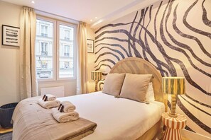 Een hotelkamer met een groot bed, een raam met gordijnen, een wand met luipaardprint en een klein rond tafeltje met een lamp.