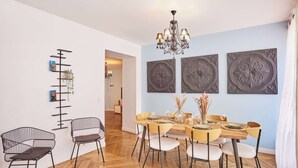 A dining area with a wooden table set for four, surrounded by chairs with white cushions. There are decorative wall panels above the table and a black metal rack with books and a plant on the wall.