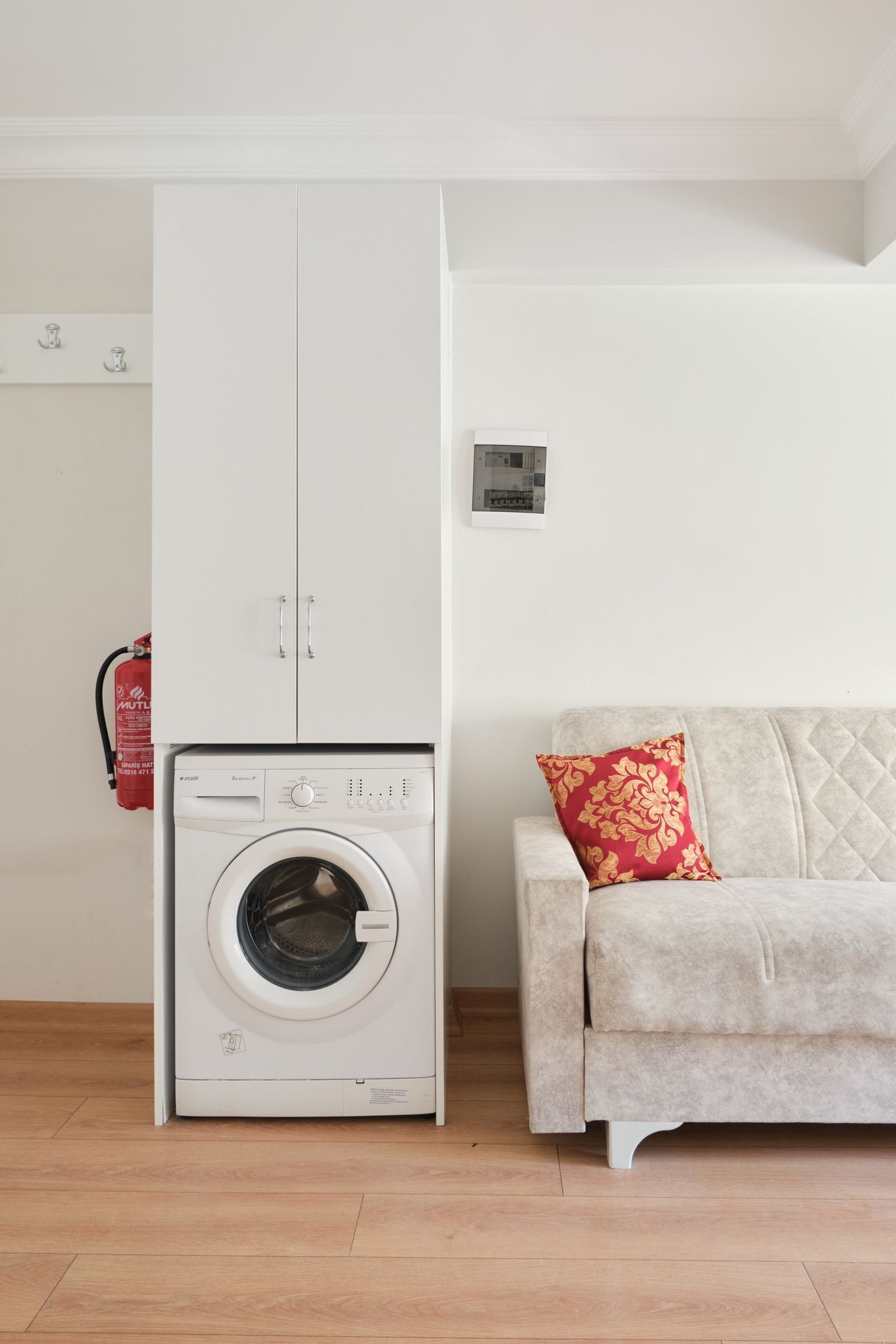A laundry area with a washing machine, a white wardrobe, and a beige sofa with a red cushion.
