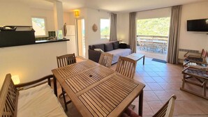 A modern room with a wooden dining table, a sofa, and a TV. There is a balcony with outdoor furniture.