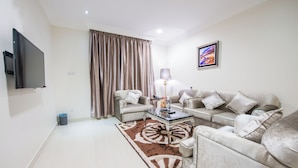 A modern living room with a flat-screen TV, two sofas, a coffee table, and a large window with curtains. - MASSET ALMASEM5 SERVICED APARTMENTS (Al-Hofuf)
