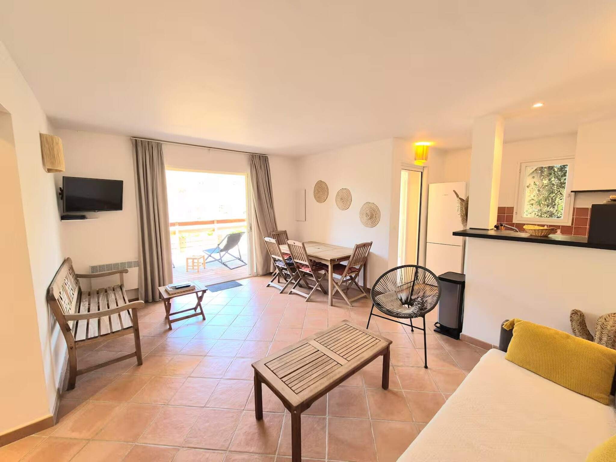 Ein geräumiges Wohnzimmer mit einer Couch, einem Couchtisch, einem an der Wand montierten Fernseher, einem Esstisch mit Stühlen und einem Balkon mit einem Sessel.