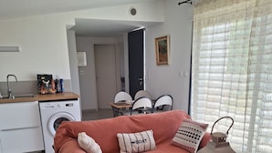 Un salon comprenant un canapé, une table basse en verre, un tapis tissé, un lave-linge et un coin repas avec des chaises.