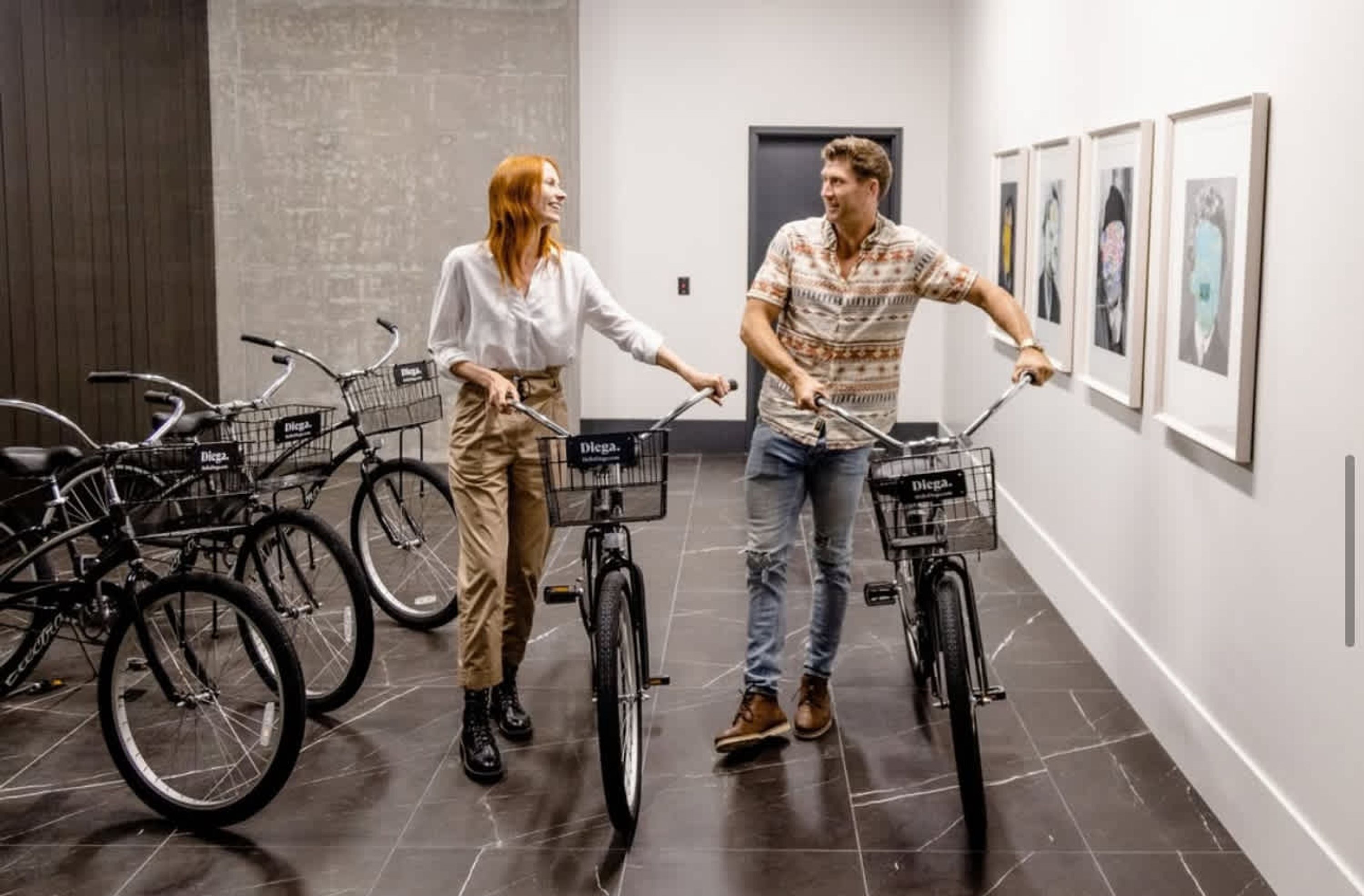 Two people on bicycles in a hallway with bicycles parked against the wall and framed artwork on the wall.