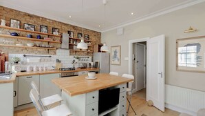 A compact kitchen with a wooden countertop, white cabinetry, and a brick wall. There are hanging lights, a refrigerator, and a dining area with chairs. - Stunning 2-bedroom Flat in Fulham (London)