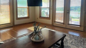 Un'ampia stanza con soffitto in legno, grandi finestre con tende, una televisione a schermo piatto montata a parete e un tavolo in legno con oggetti decorativi.