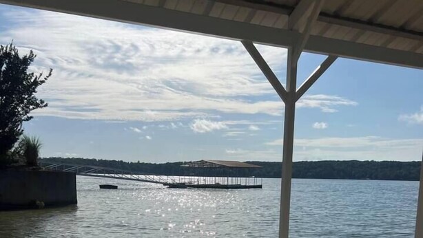 Un pontile di legno con anelli metallici, affacciato su un lago calmo con un molo e alberi in lontananza.
