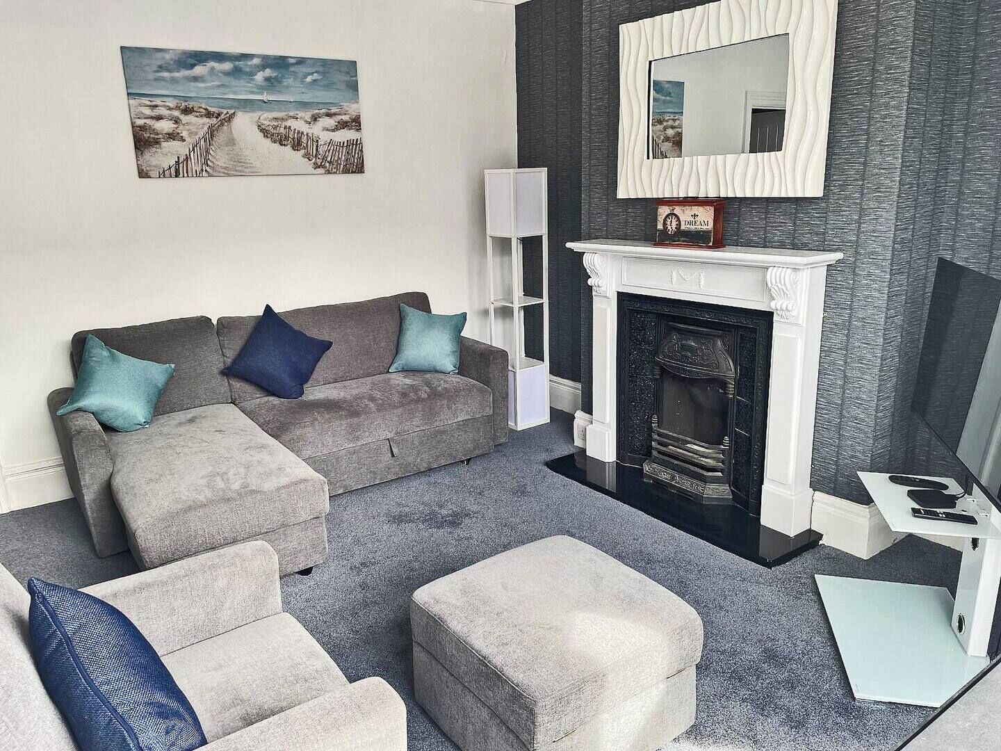 Una sala de estar con un sofá gris, dos cojines azules, una mesita auxiliar blanca, un televisor de pantalla plana y una chimenea.