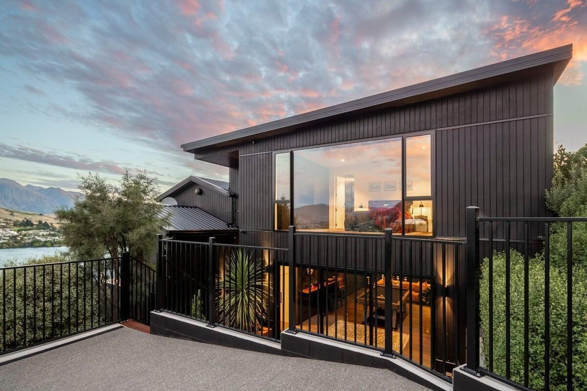 Een modern huis met grote ramen, een balkon en uitzicht op een meer en bergen.